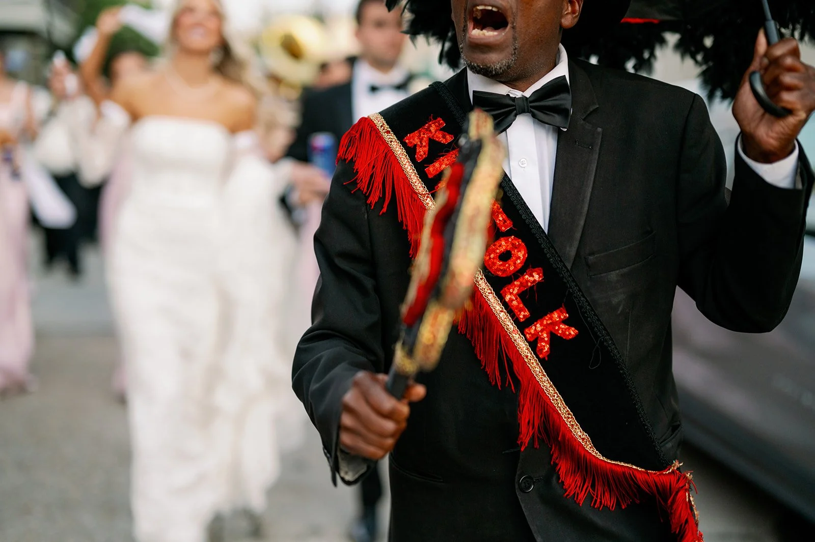 A man in a black tuxedo and bow tie carrying a decorative baton, wearing a red and black sash that says 'HOK' and '2023', leading a parade of people dressed in white gowns and formal attire, with blurred crowd in the background.