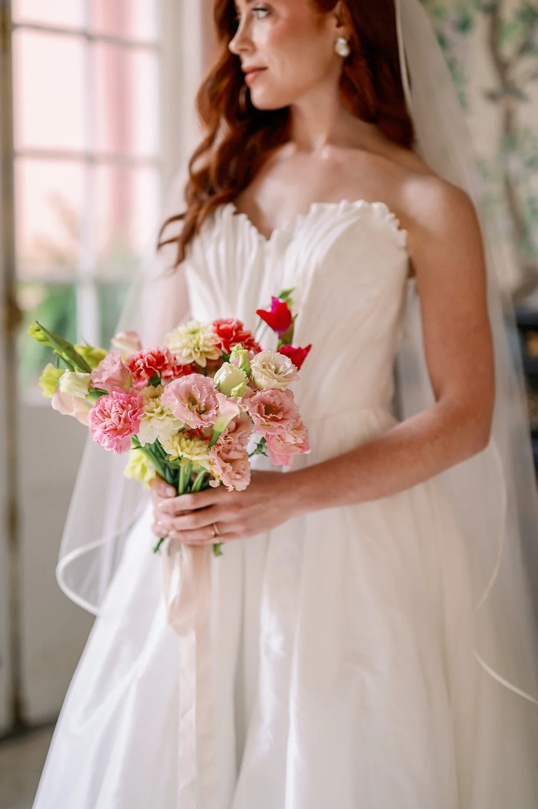 A bride in a white wedding dress holding a bouquet of pink, red, and cream flowers, standing indoors near a window.