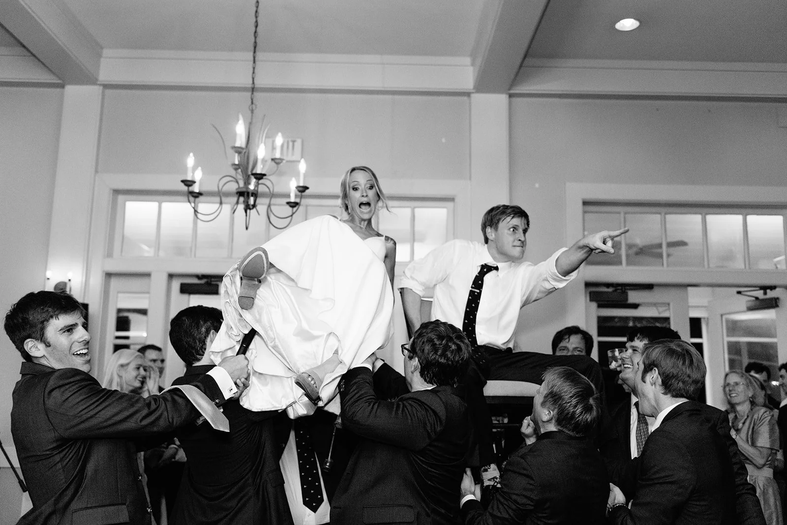 A wedding reception with people lifting a bride and groom in chairs. The bride looks surprised, and the groom is pointing forward. The reception is indoors with a chandelier and large windows.