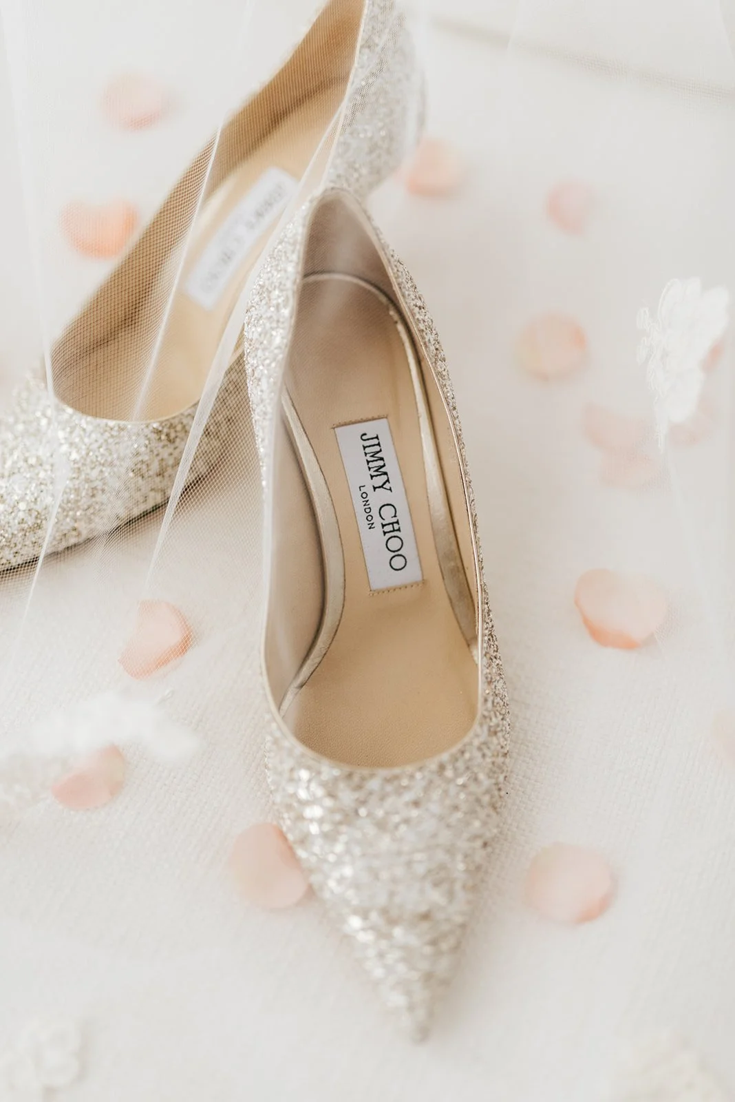 A pair of sparkling gold Jimmy Choo high-heeled shoes on a white surface decorated with pink flower petals.