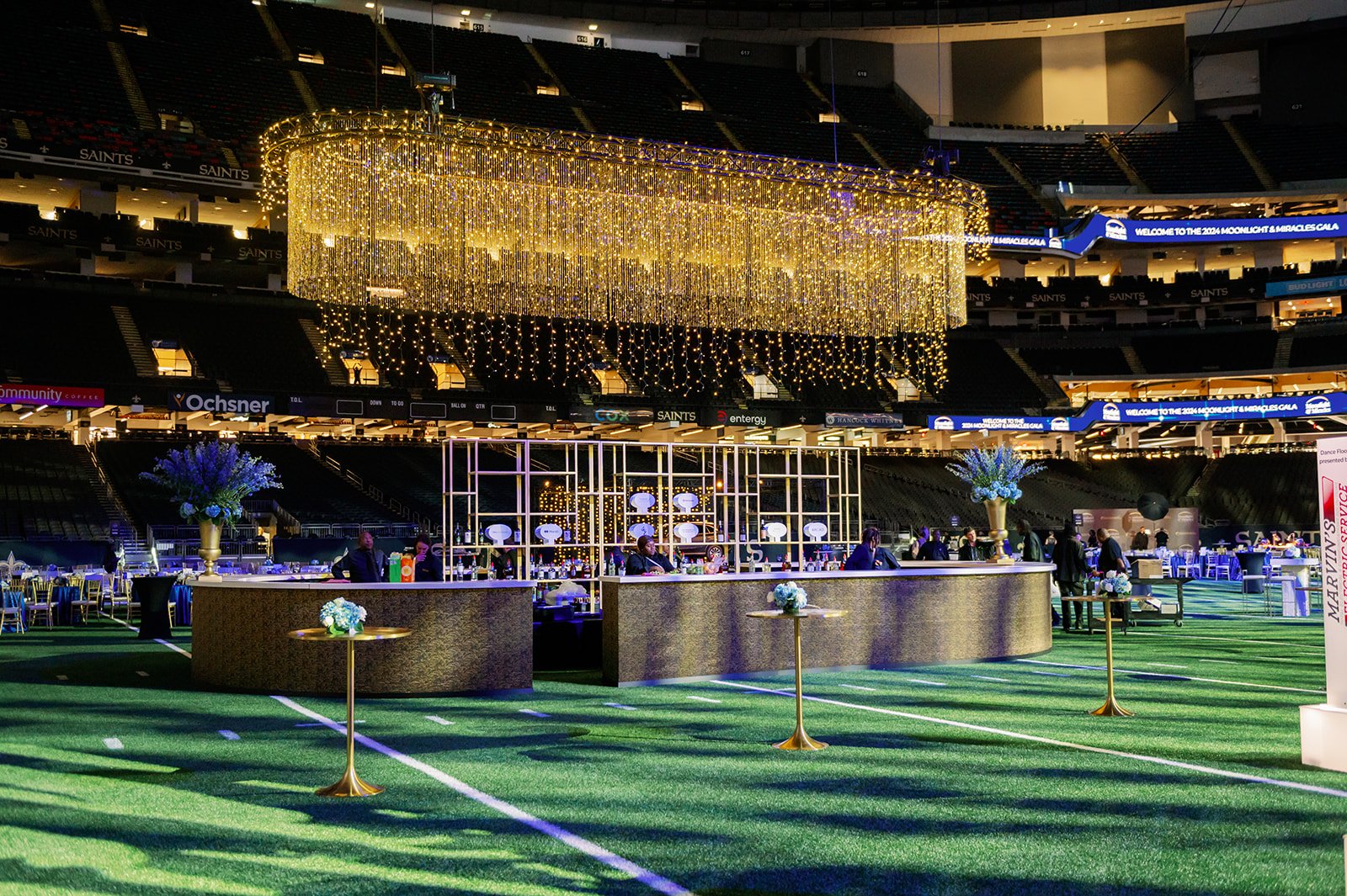 A decorated indoor event space set up on a sports field with artificial turf. The area features a large, circular chandelier with golden hanging lights, floral arrangements, a bar with staff, and tables with floral centerpieces. In the background, th
