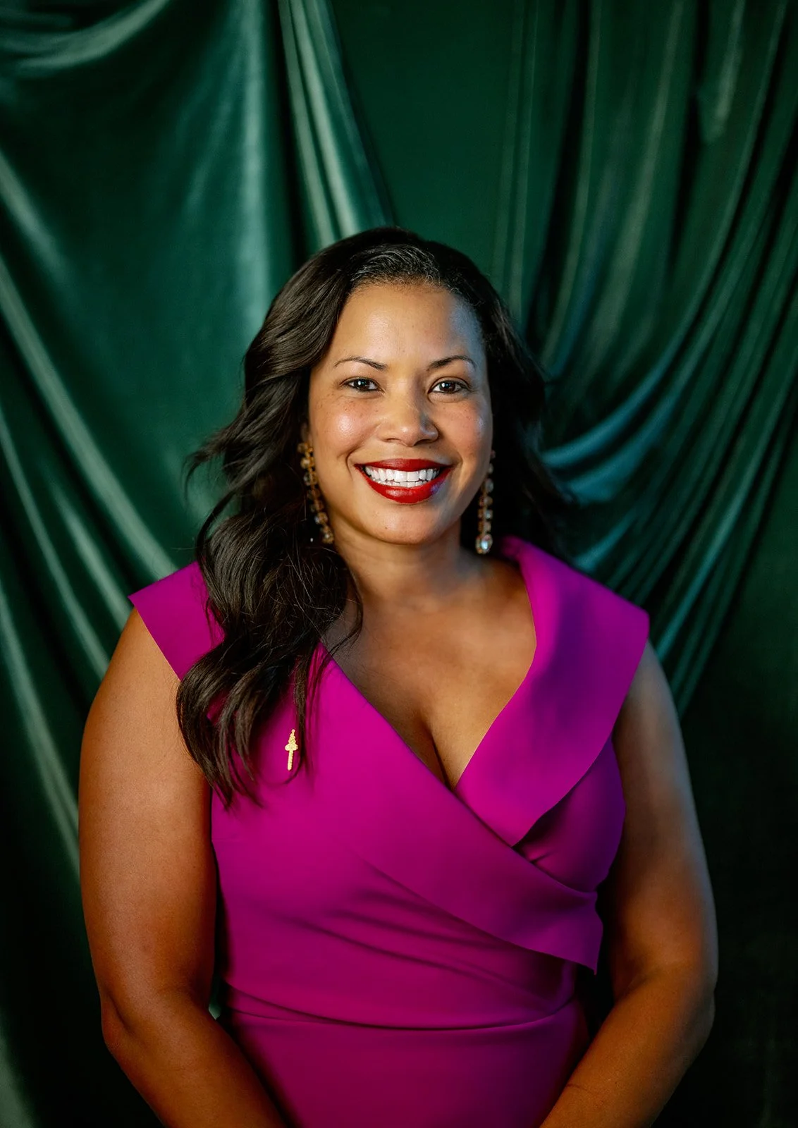 A woman smiling in a bright pink dress with pearl earrings, standing in front of green draped fabric.