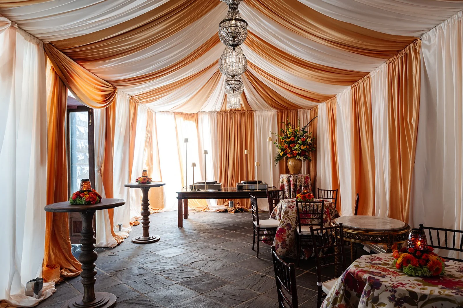 Elegant interior decorated with orange and cream drapes, floral arrangements, and chandeliers, set up for an event or celebration.