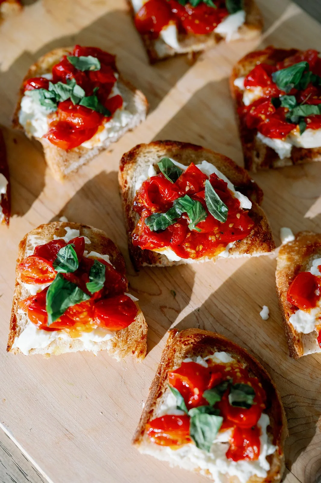 Close-up of several pieces of toasted bread topped with tomato sauce, fresh basil, and dollops of white cheese on a wooden surface.