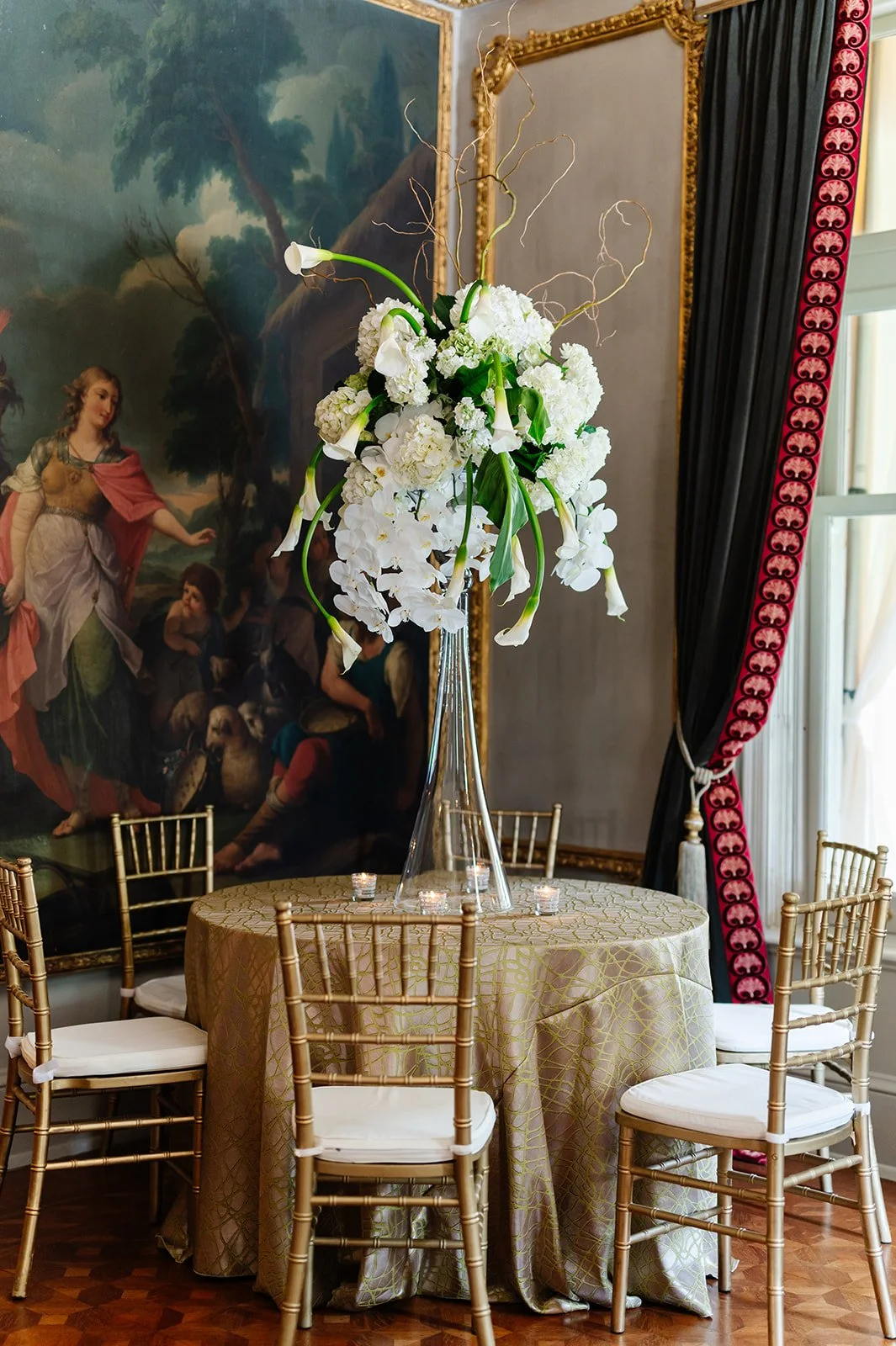 Elegant floral arrangement of white flowers in a tall glass vase on a round table draped with gold patterned tablecloth, surrounded by gold chairs with white cushions, in a room with large oil painting and dark curtains.