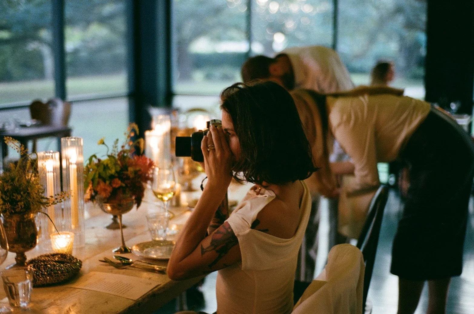 A young woman taking a photo with a camera at a decorated table during a social gathering, with two people in the background near large windows showing greenery outside.