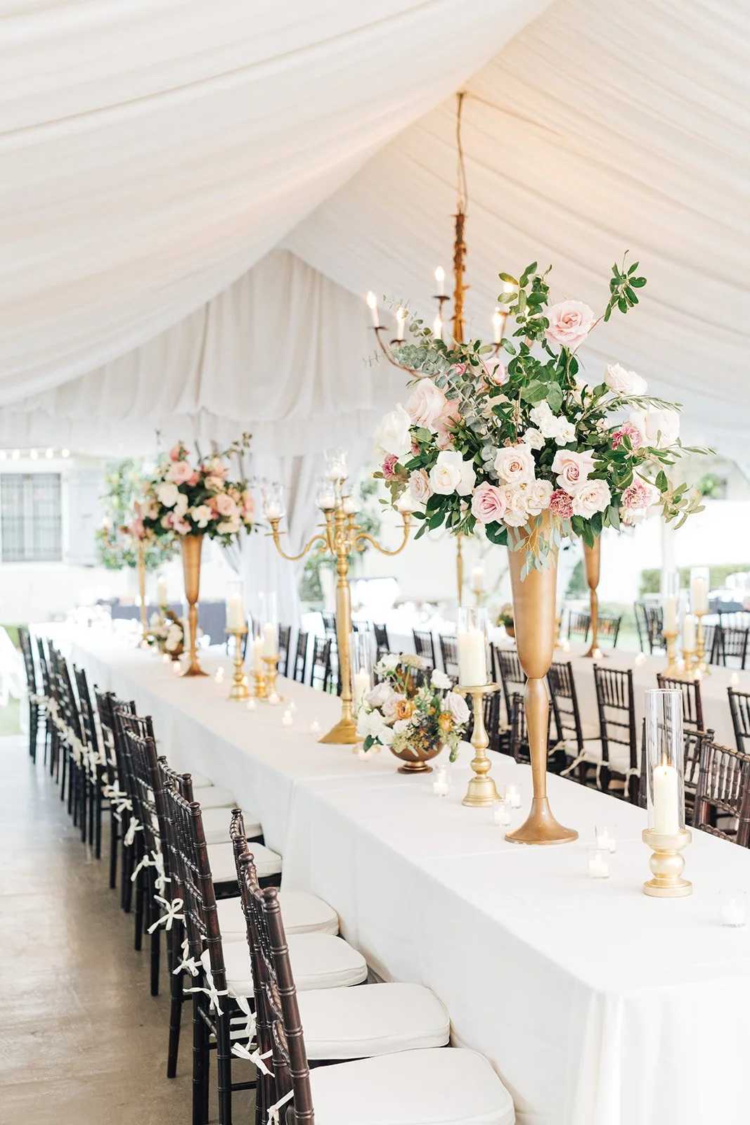 Elegant wedding reception table decorated with tall floral centerpieces, gold candelabras, and candles under a white canopy tent.