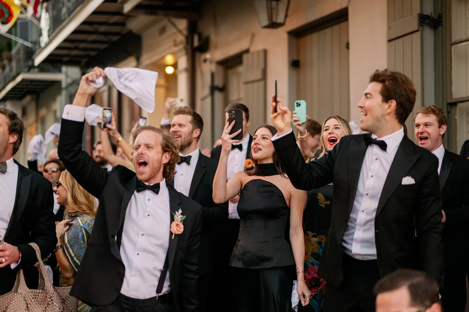 A group of people dressed in formal black tie attire, smiling and taking photos at an outdoor event, possibly a wedding or celebration, with a building in the background.
