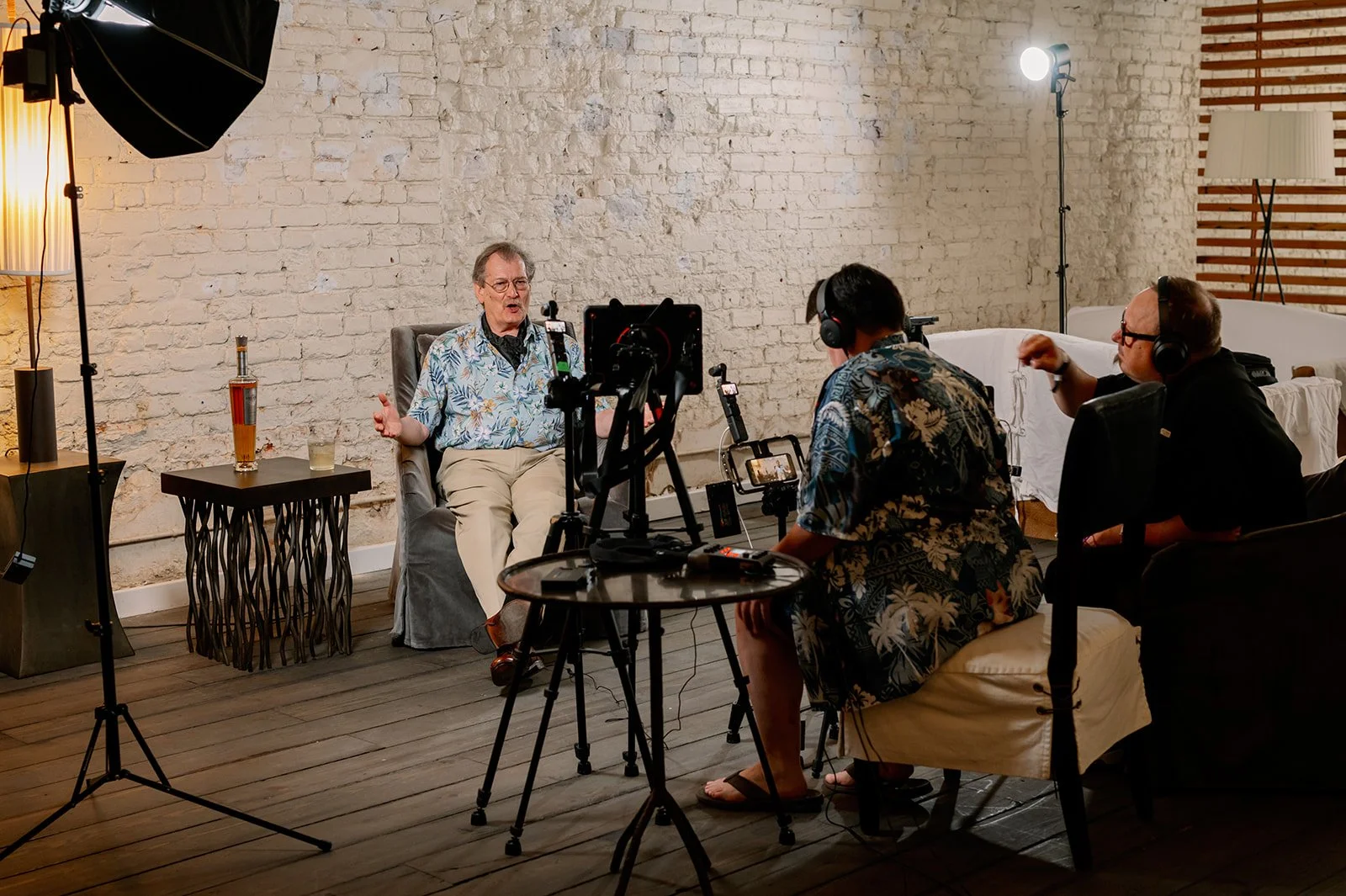 A man in a floral shirt being interviewed in a studio with a brick wall background, with two crew members recording and filming him.