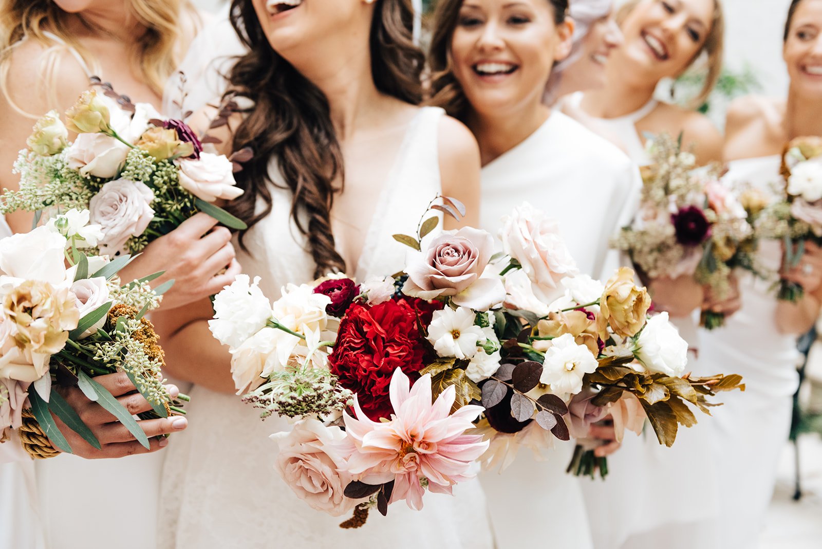 A bride and bridesmaids holding bouquets of mixed flowers, including roses and dahlias, at a wedding celebration.
