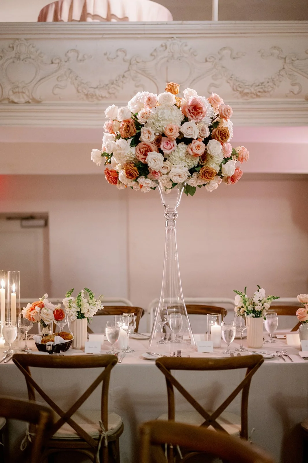 Elegant banquet table decorated with a tall floral centerpiece of pink, white, and light brown roses, surrounded by smaller floral arrangements, candles, glasses, and place settings in a well-lit, ornate room.