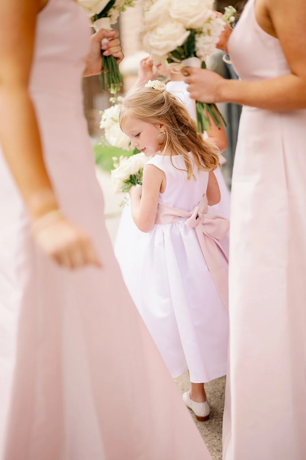 A young girl in a pink dress with a bow in her hair holding a bouquet of white flowers at a wedding, surrounded by women in pale pink dresses holding similar bouquets.