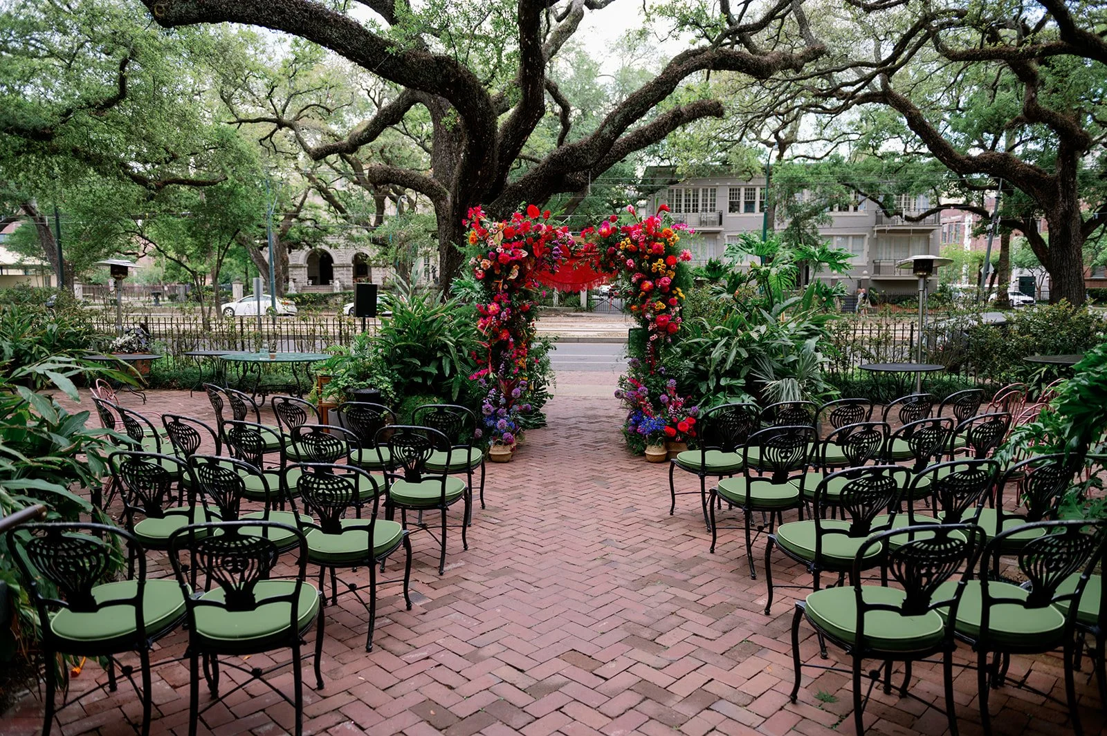 Outdoor wedding ceremony setup with black chairs with green cushions facing a floral arch in a garden, under large leafy trees.