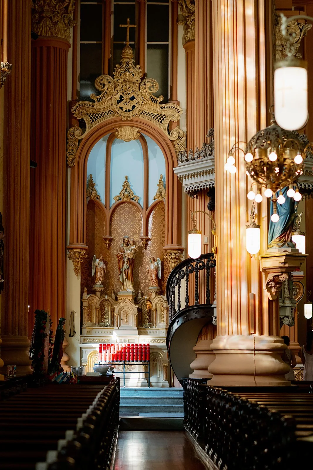 Interior of a church with an ornate altar displaying statues of religious figures, lit candles, and decorative woodwork.