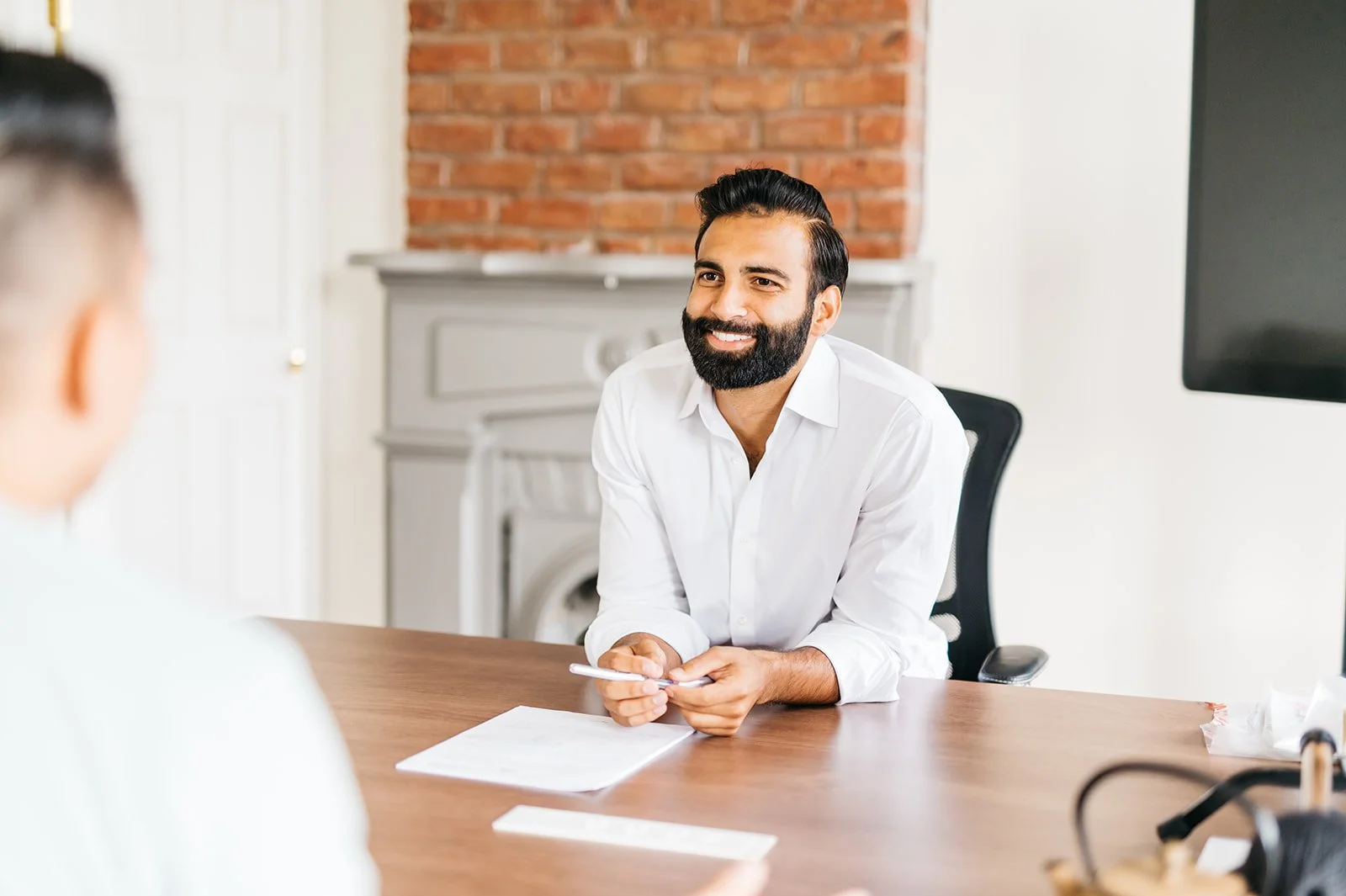 A bearded man in a white shirt smiling and sitting at a desk during a conversation in an office with white walls, a television, and a brick fireplace in the background.
