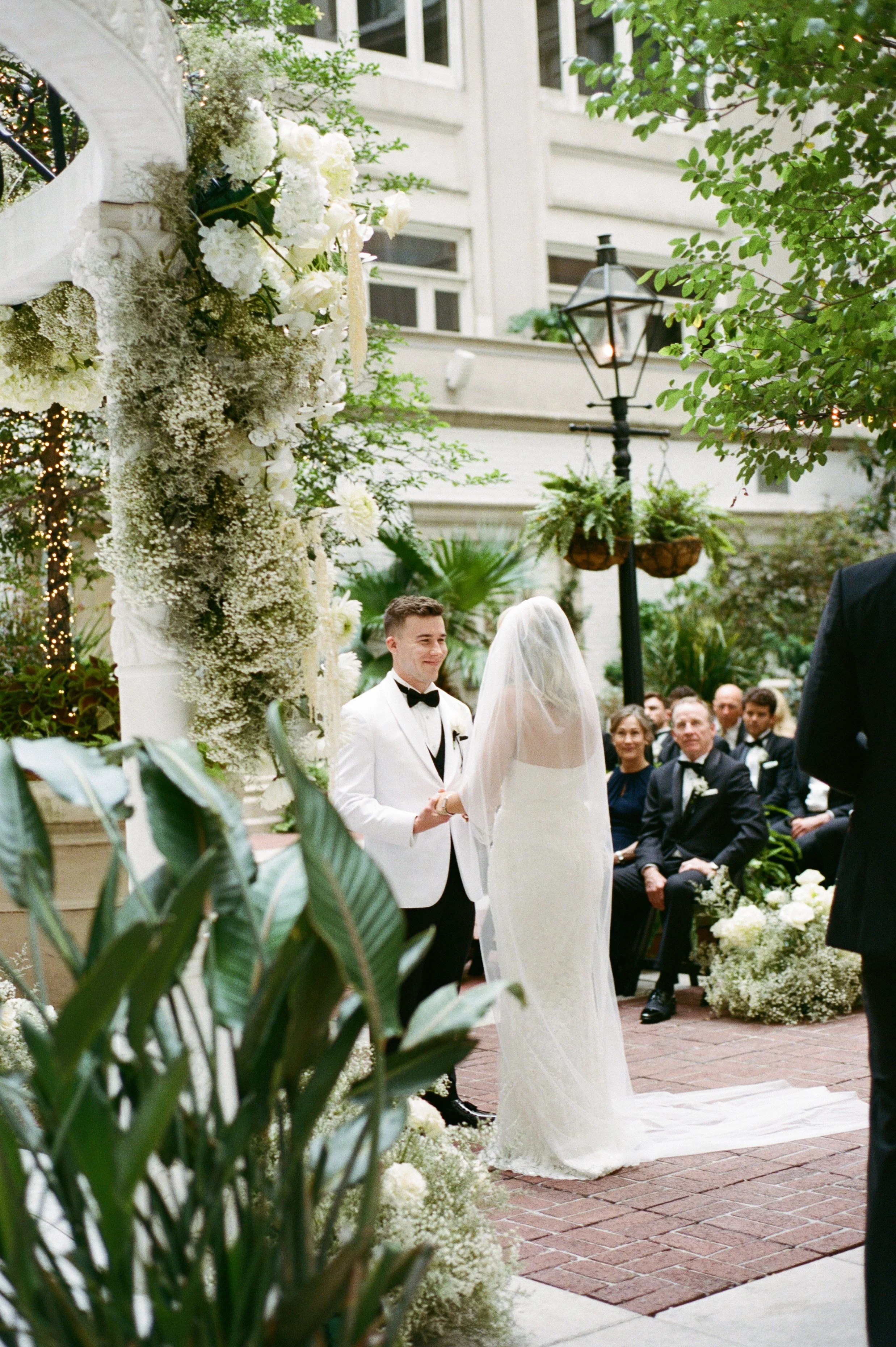 A bride and groom exchanging vows during a wedding ceremony outdoors, surrounded by floral decorations and seated guests, with a white building and trees in the background.