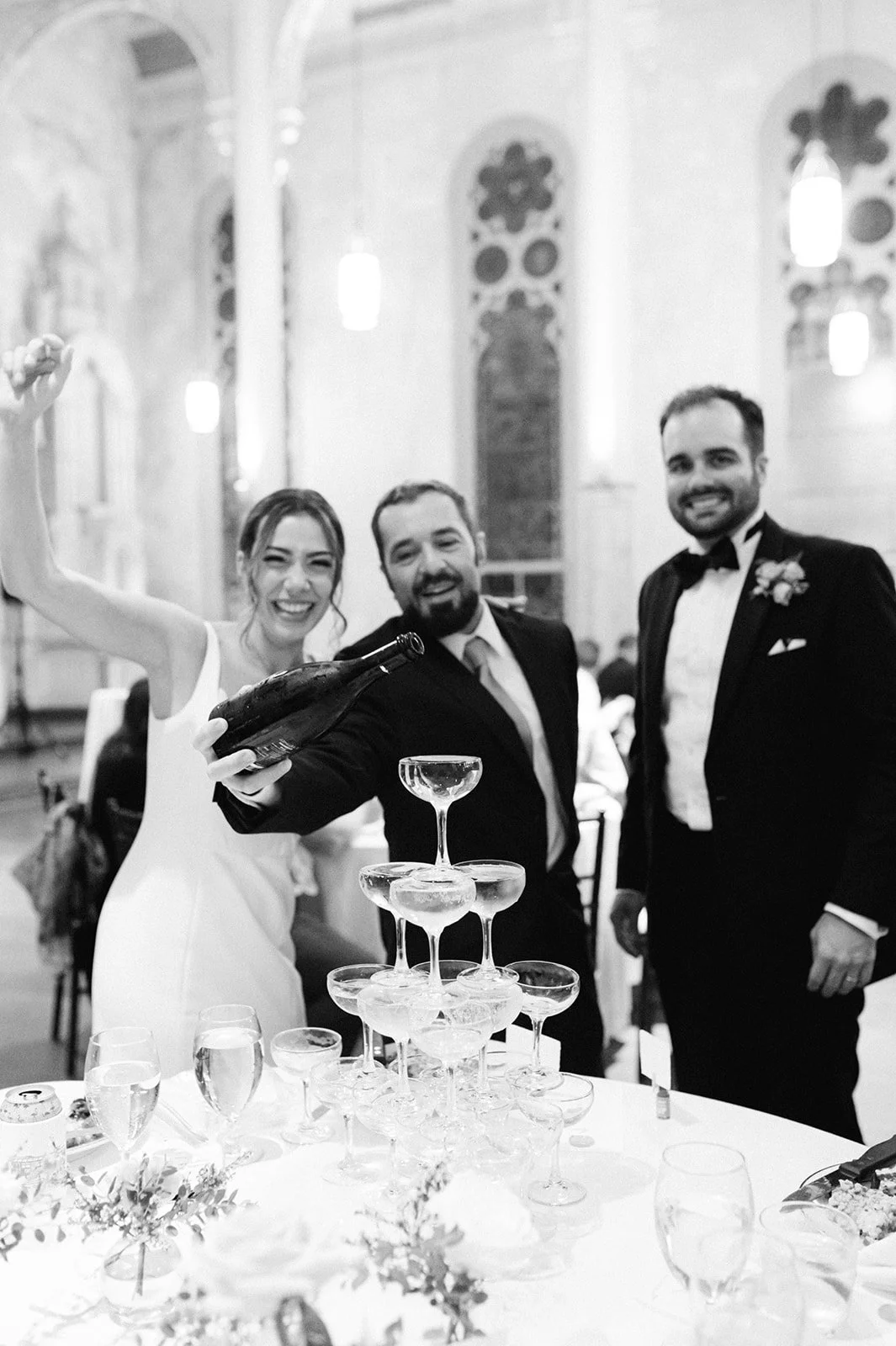 Bride, groom, and a guest at a wedding celebration pouring champagne into a pyramid of glasses in a church.