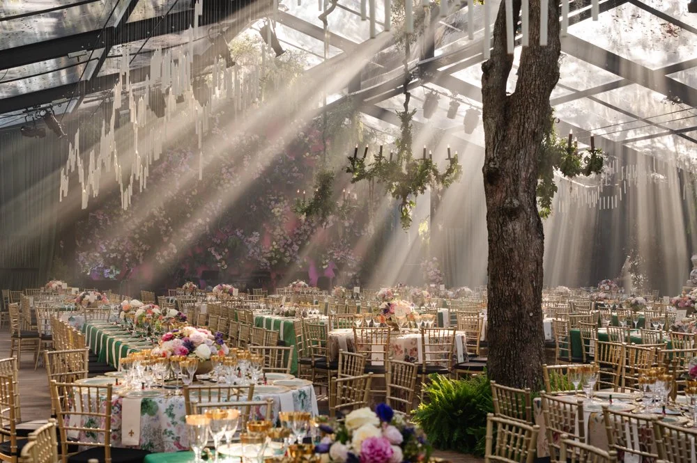 Sunlight beams through a glass-ceilinged event space illuminating tables set for a celebration, with floral centerpieces and gold chairs.