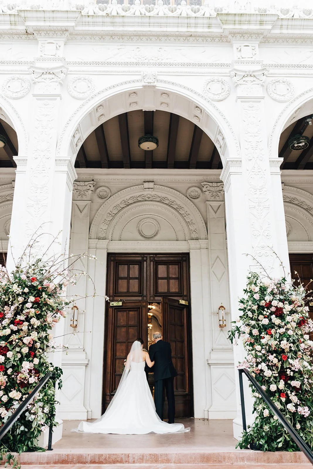 A bride and an older man, likely her father, standing at the entrance of a grand, ornate building. The woman is in a white wedding gown with a veil, and both are facing away from the camera. The entrance is decorated with large floral arrangements on