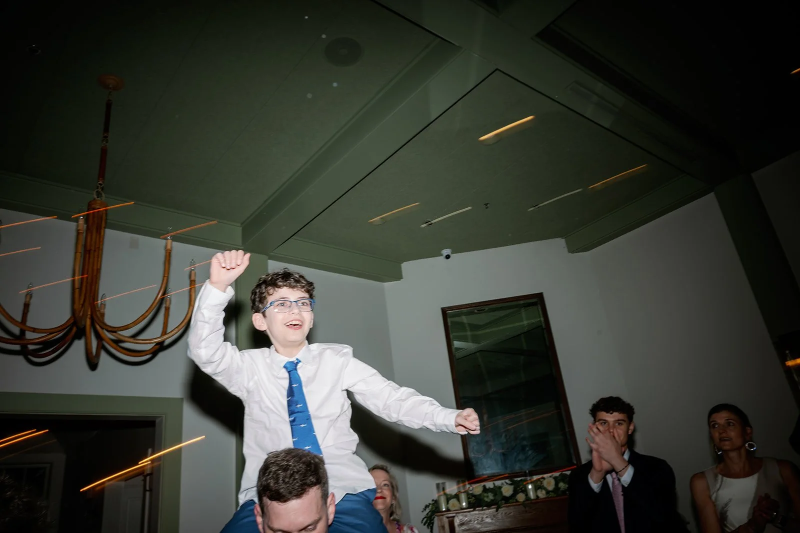 A boy with glasses and a blue tie sitting on someone's shoulders, celebrating at an indoor event, with other well-dressed people clapping and smiling in the background.