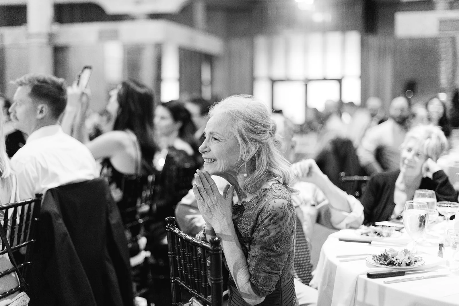 An elderly woman smiling and clapping at a formal event or banquet, seated at a table with others in an elegant setting.