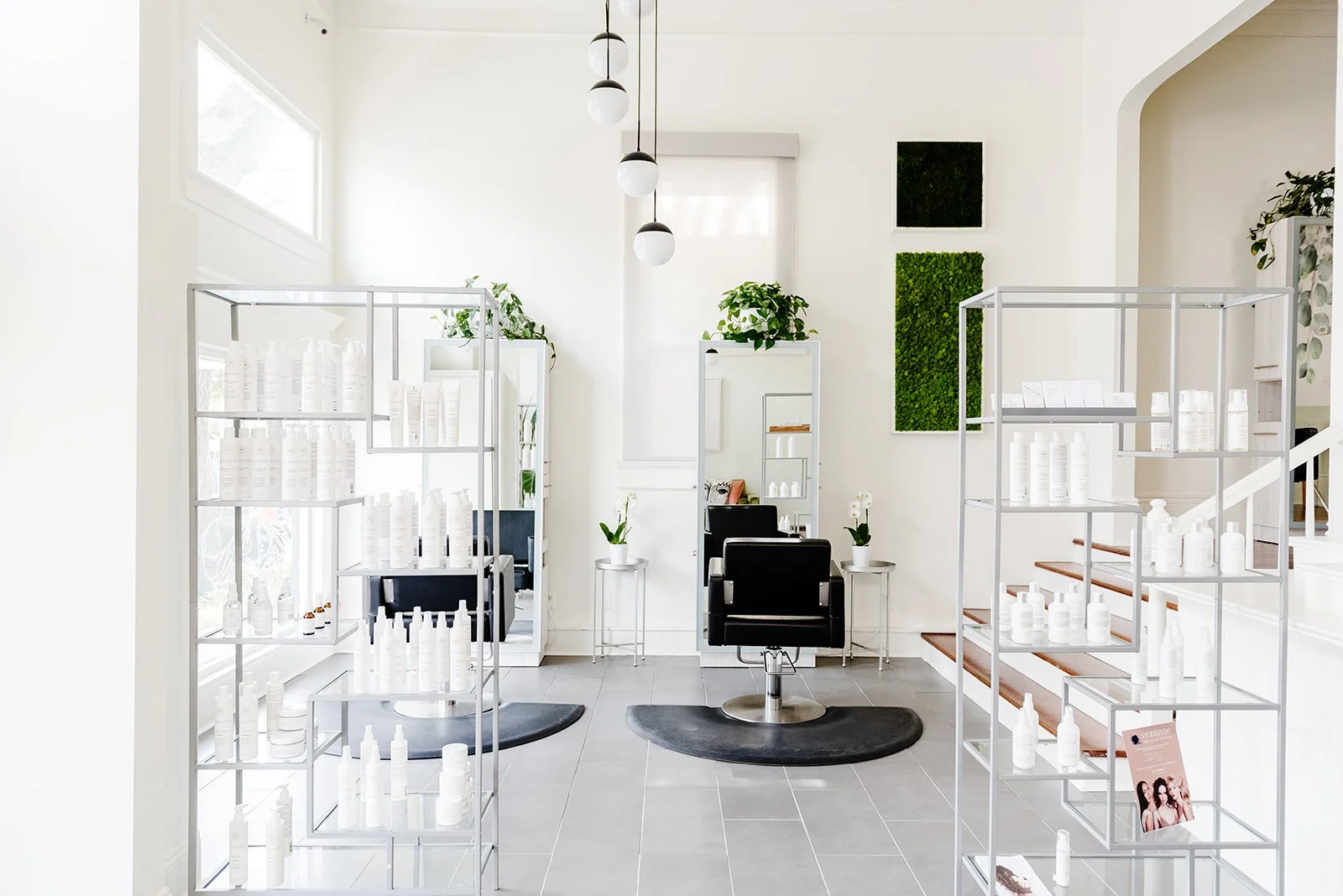 A modern and minimalistic hair salon or beauty studio with black salon chair, mirror, potted plants, white shelves with bottles, and staircase with magazines.
