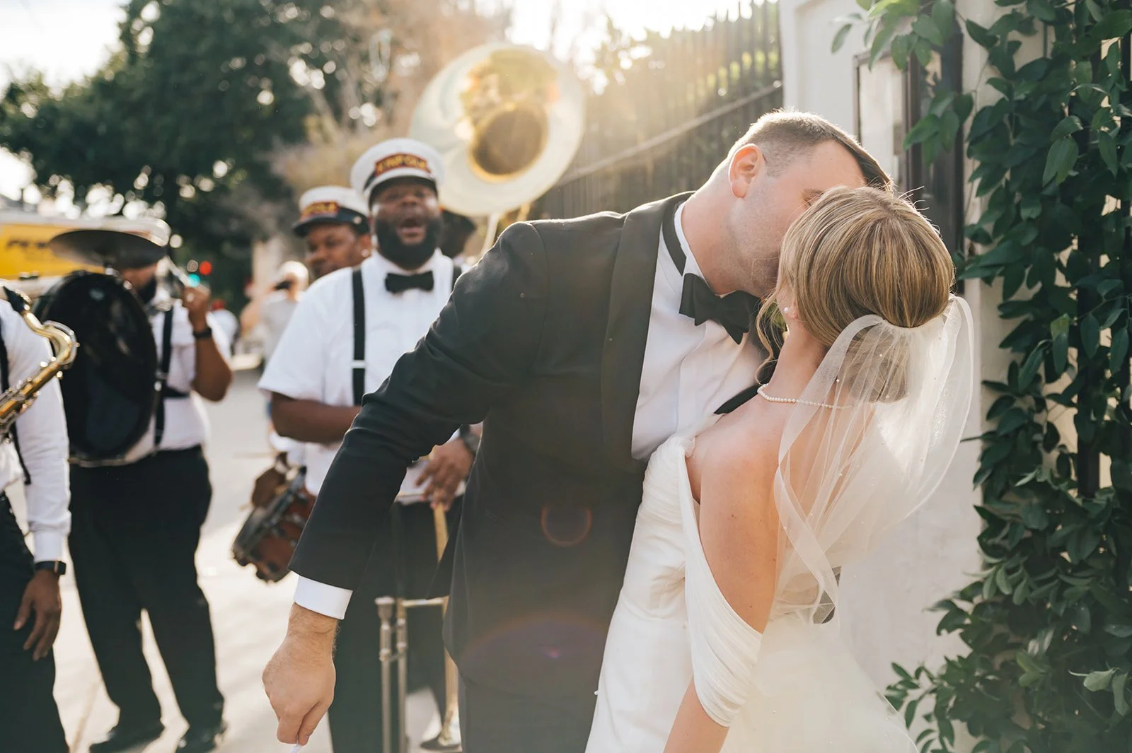 A newlywed couple sharing a kiss outdoors, surrounded by a brass band playing music, on a sunny day.