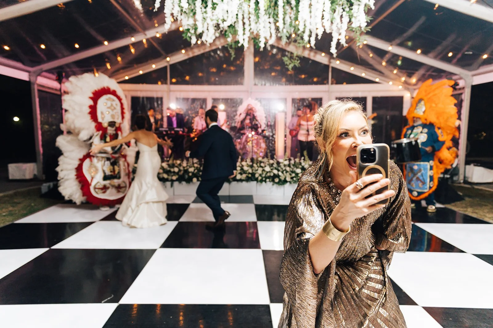 A woman in a shiny bronze dress is taking a selfie at a wedding reception, with a dance floor, brides and grooms dancing, and performers with colorful costumes and feathered headdresses in the background.