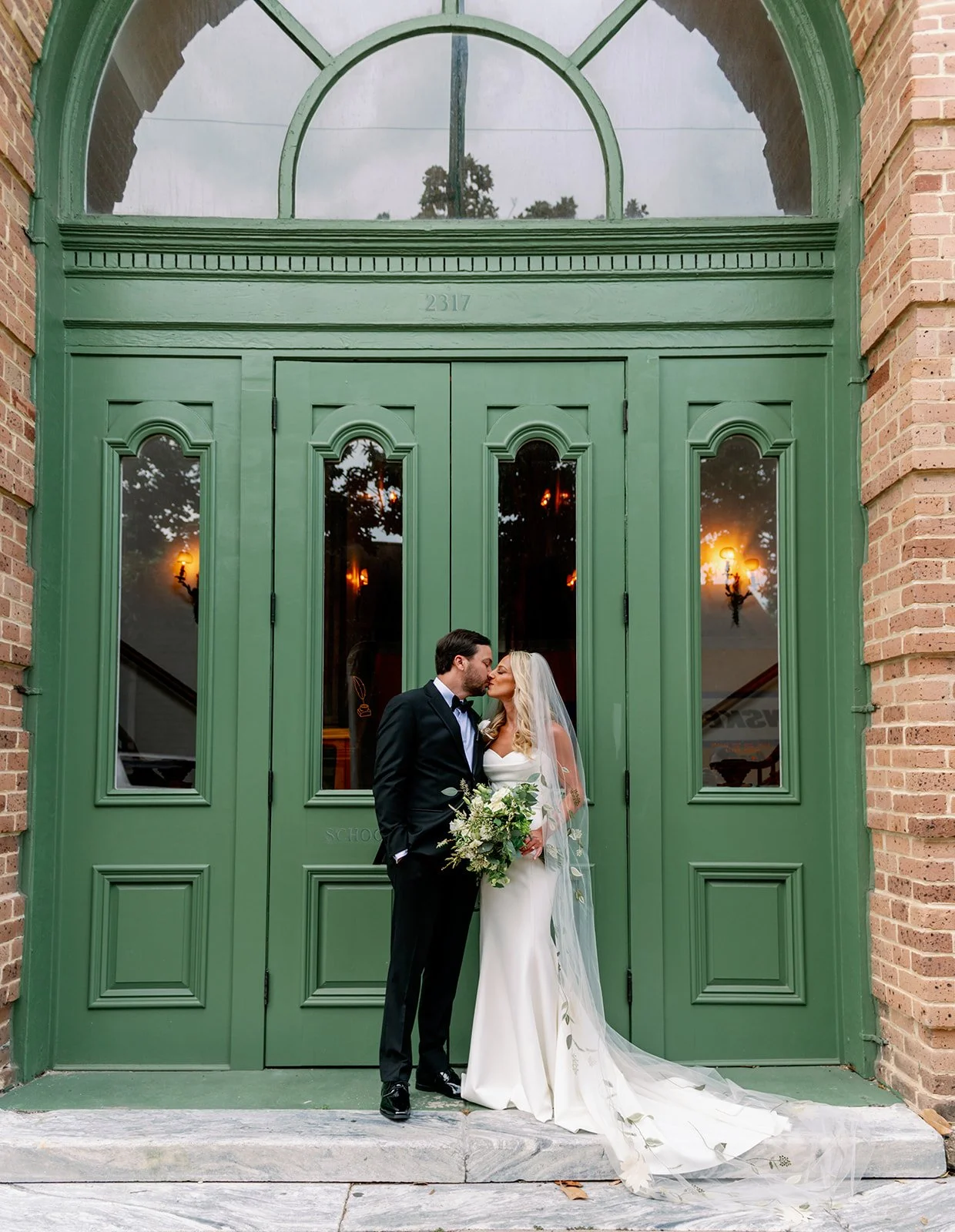 Bride and groom standing close, about to kiss in front of a large green door with glass panes, outside a brick building, at sunset.