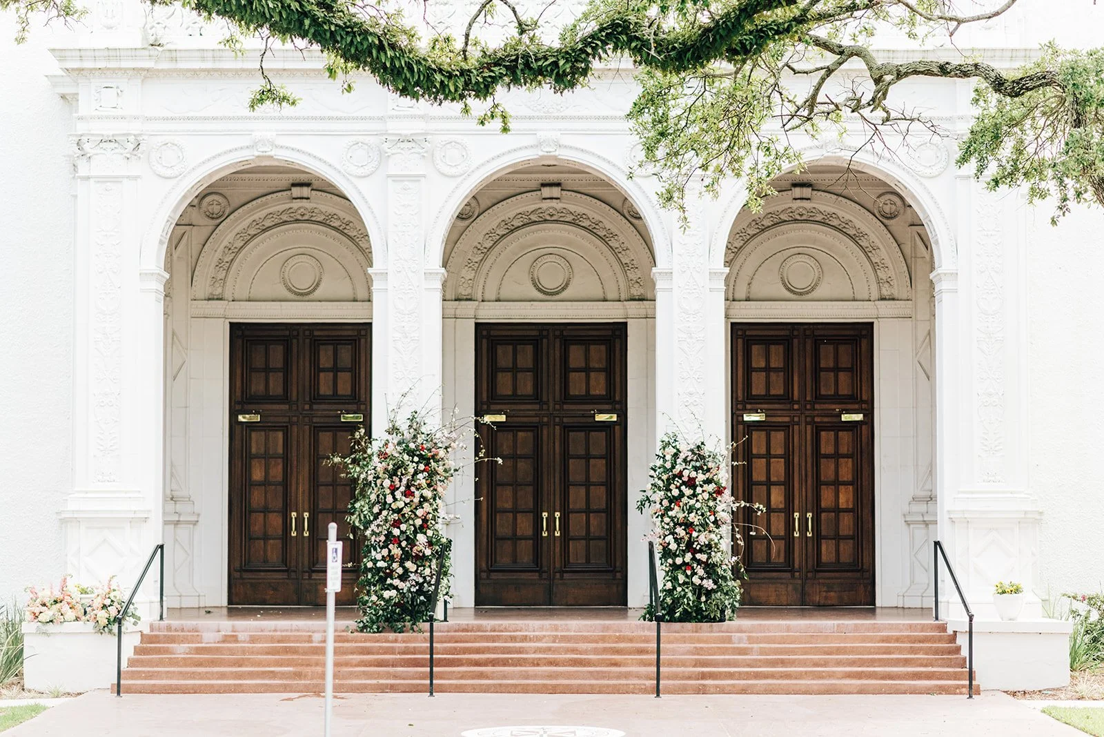 A church entrance with three large dark wood doors, surrounded by white architectural details and decorated with floral arrangements.