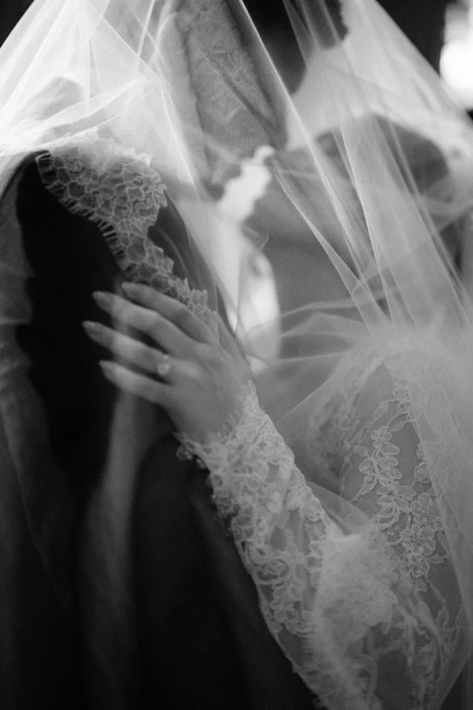 Black and white photograph of a bride behind a sheer veil, wearing lace gloves and holding her hand to her face, with a wedding ring visible.