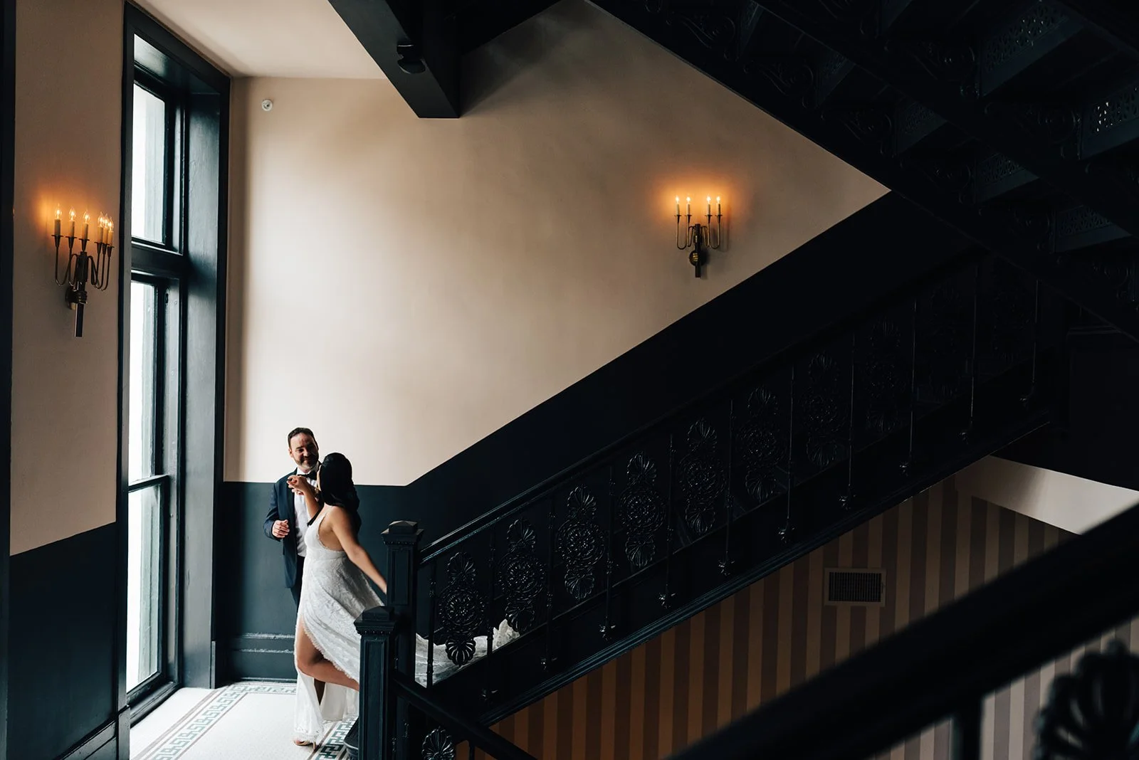 A bride in a white wedding dress and a groom in a dark suit dance on a staircase inside a dimly lit elegant venue with large windows and wall-mounted candle-style lights.
