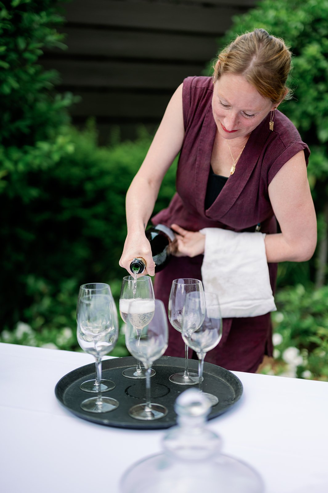 A woman with red hair, wearing a sleeveless maroon dress, is pouring champagne into a flute at an outdoor table with empty glasses, greenery in the background.
