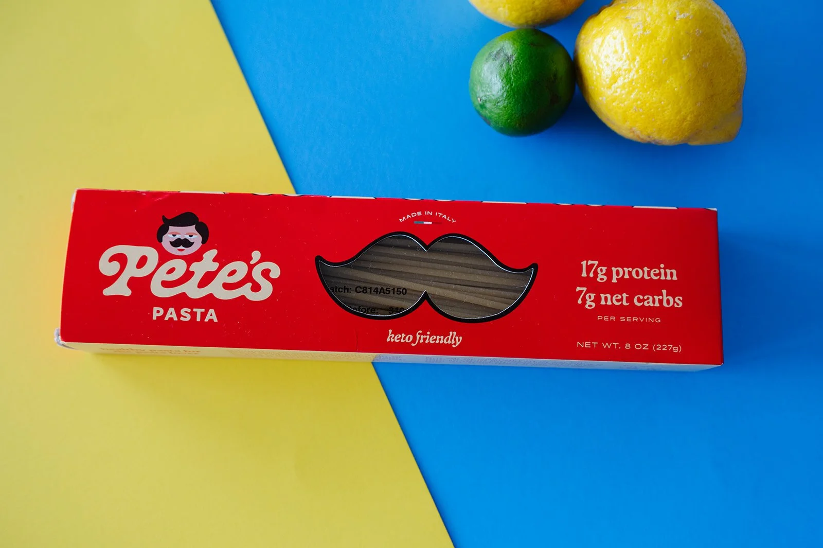 Red box of Pete's Pasta with a mustache logo and cutout window shaped like a mustache, placed on a yellow and blue background with lemons and limes nearby.