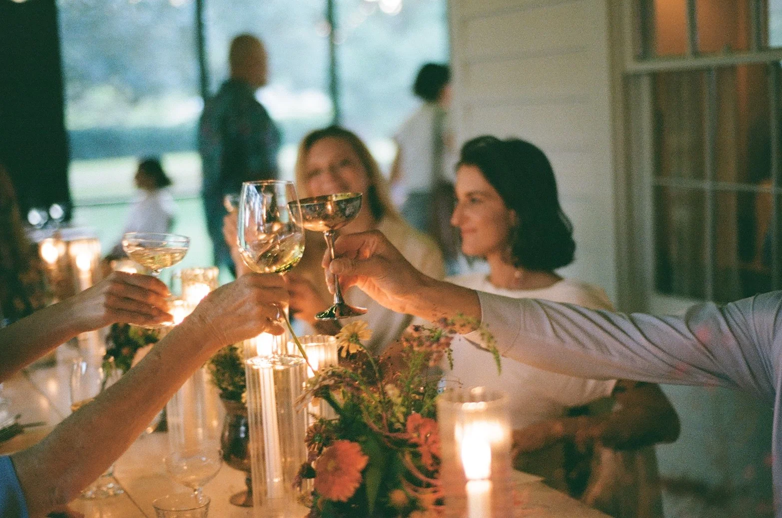 People raising glasses in a toast at a celebration, seated around a decorated table with candles and floral arrangements.