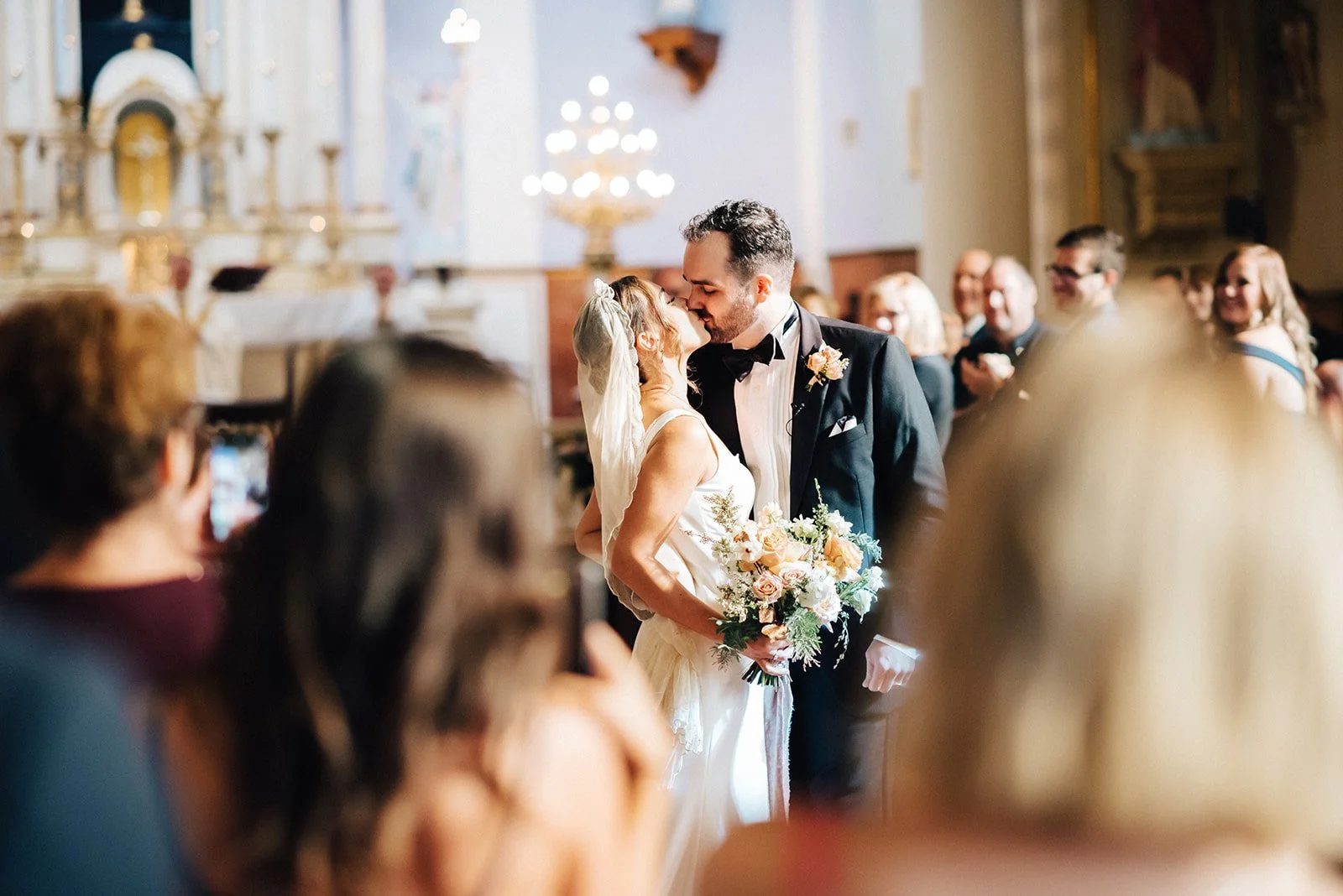 A bride and groom share a kiss during their wedding ceremony inside a church. The bride holds a bouquet of flowers, and guests watch and smile in the background.