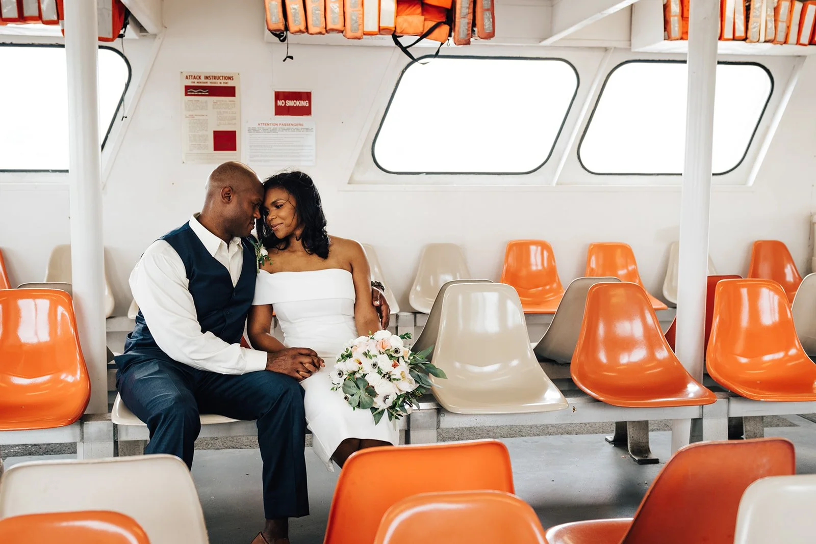 A couple dressed in wedding attire sitting closely on a row of beige and orange seats inside a ferry, with the woman holding a bouquet of flowers.