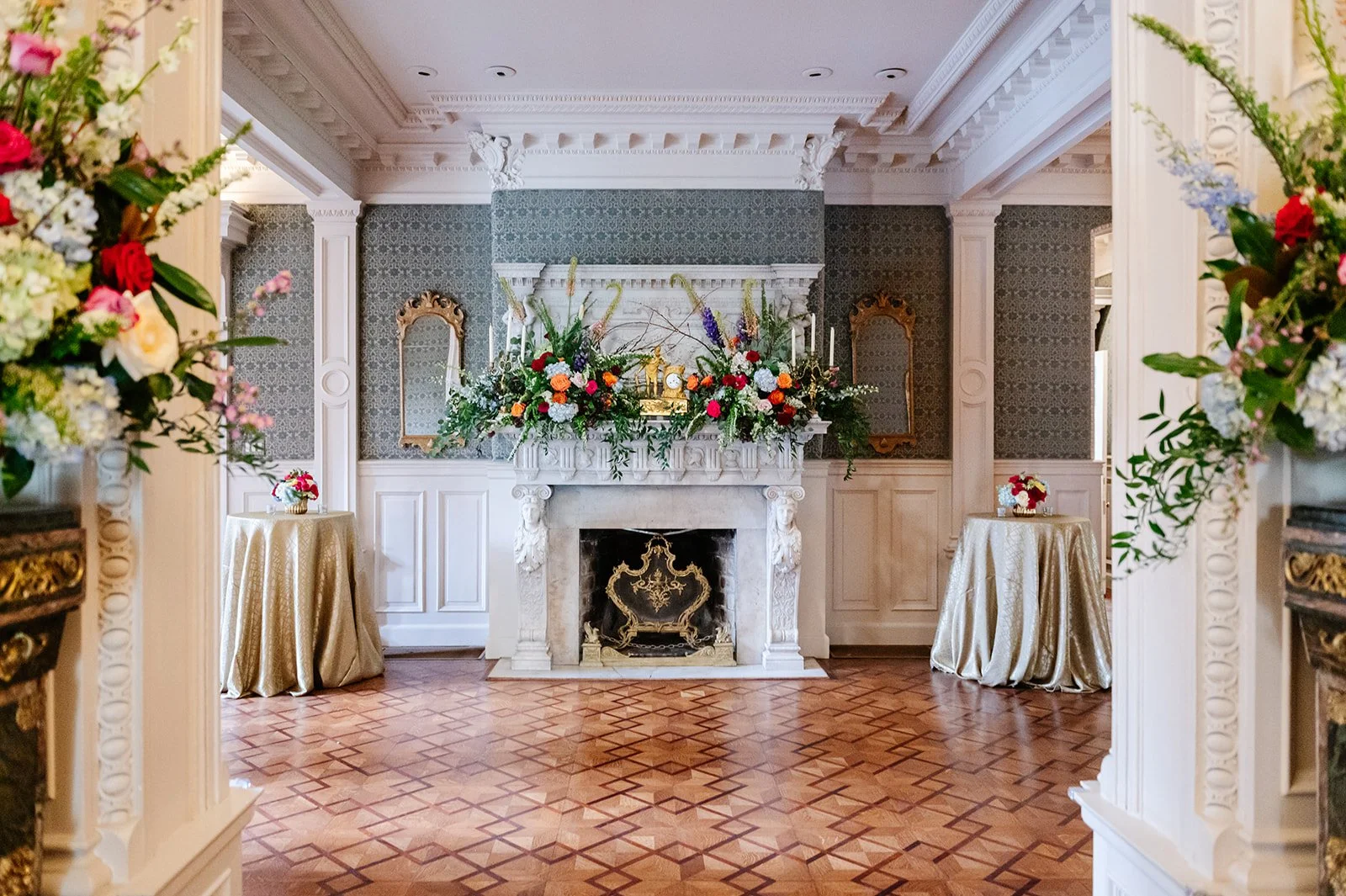 Elegant room with a marble fireplace decorated with floral arrangements and golden clocks, patterned wallpaper, and gold-framed mirrors, with two gold tables with floral centerpieces on either side.