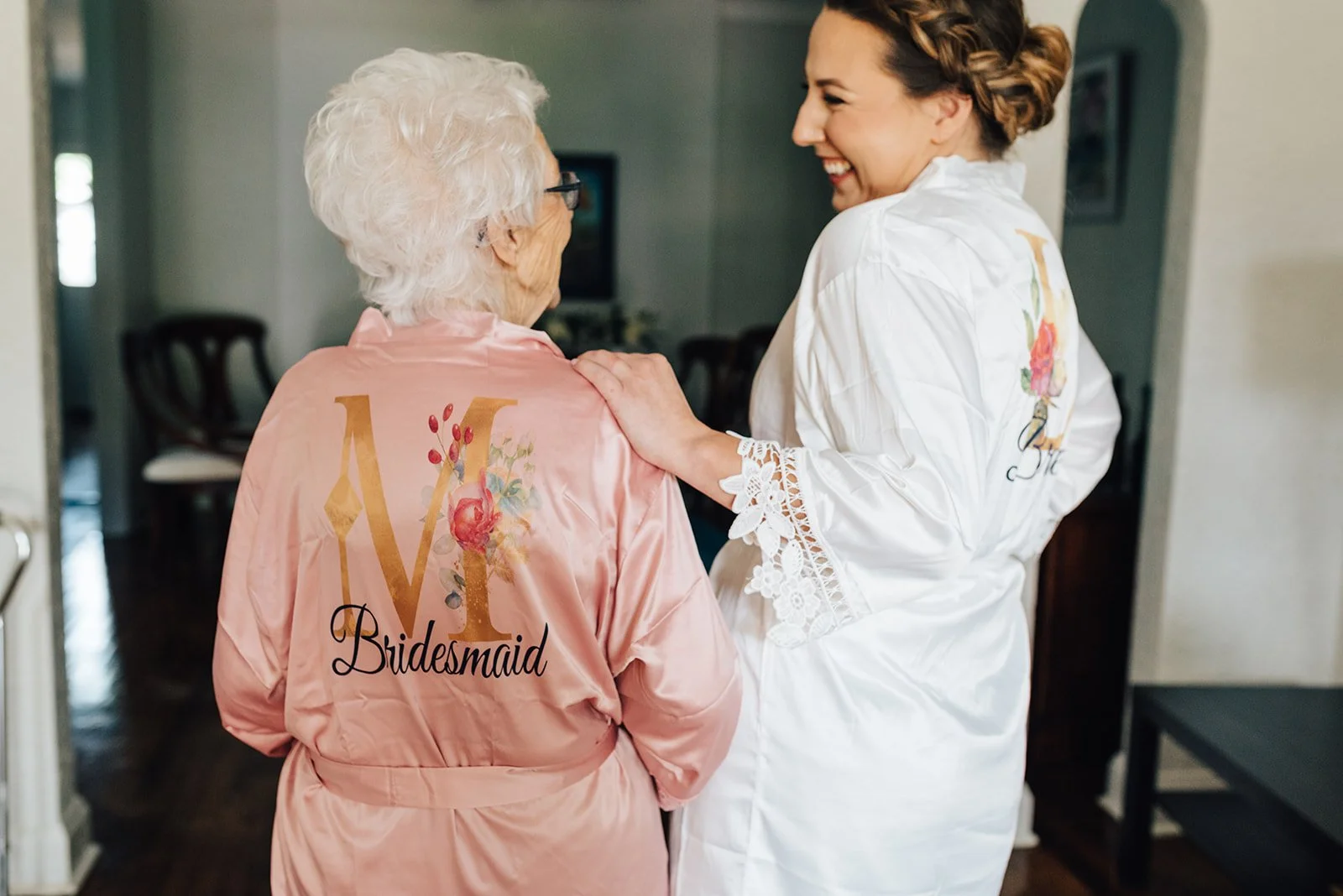 A bride and an older woman with white hair, both in satin robes with floral designs and the words 'Bridesmaid' and 'M' on the back, sharing a happy moment.