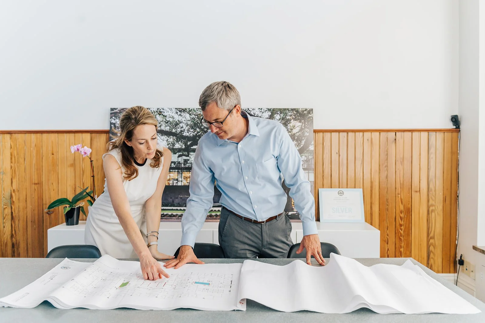 A woman and a man discussing architectural plans at a table in an office, with framed certificates and a photo of a park in the background.