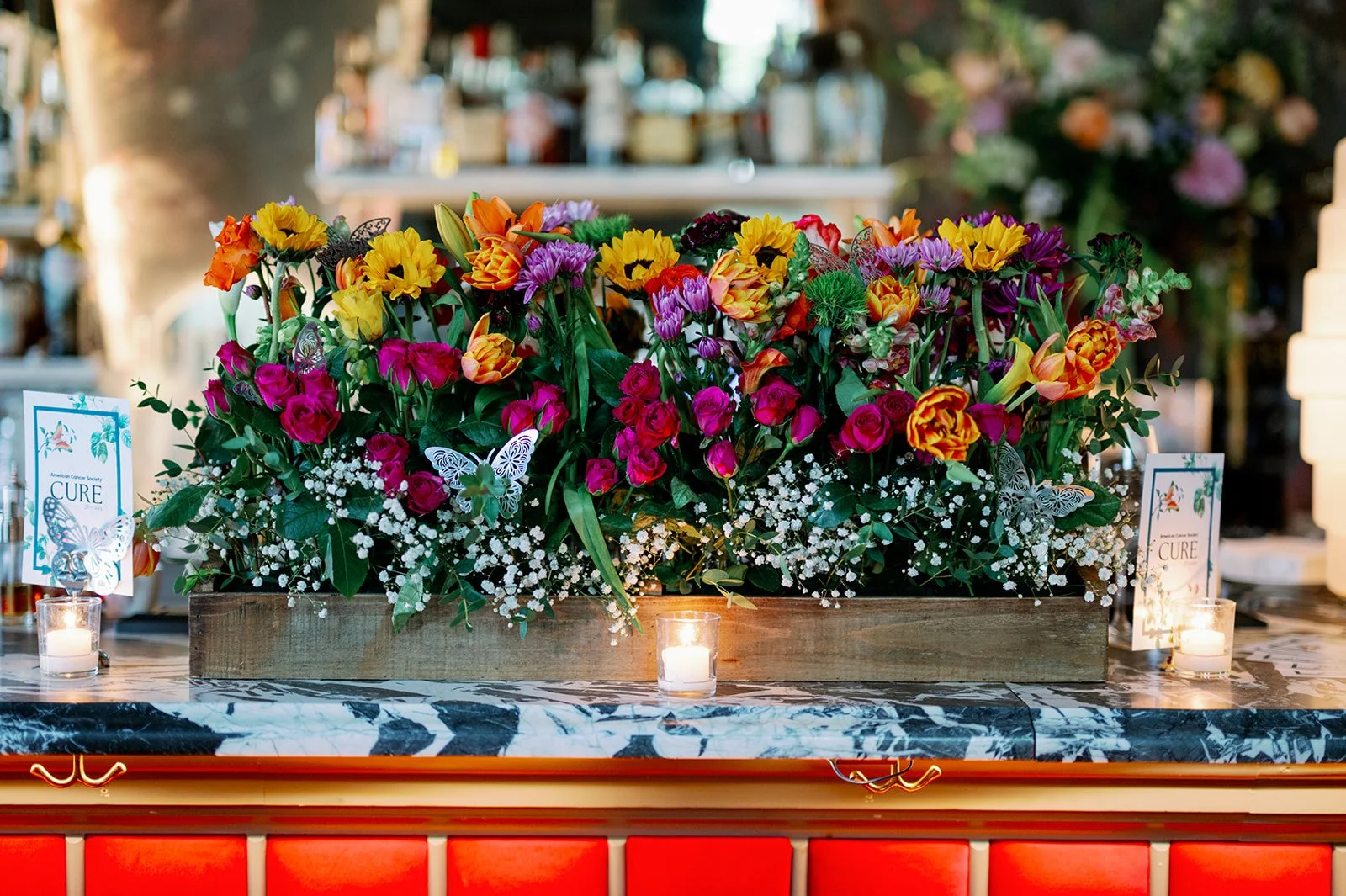A colorful floral arrangement with pink, yellow, orange, purple, and white flowers on a marble-topped table. Small candles and informational cards are placed around the flowers, with a blurred background of a bar or cafe interior.