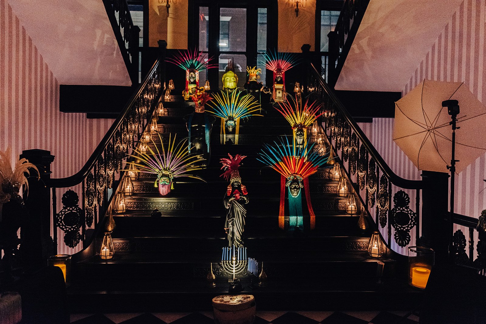 Colorful decorative masks with feathered headdresses arranged on a staircase in a dimly lit indoor setting, with lanterns and decorations on the sides.