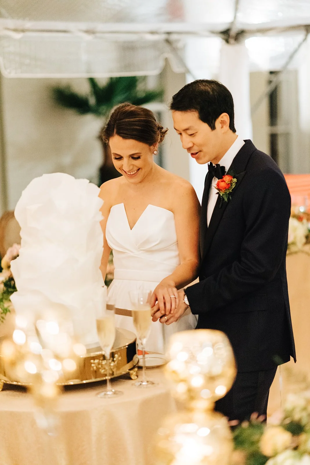 A bride and groom cut a wedding cake together at their reception, smiling happily.
