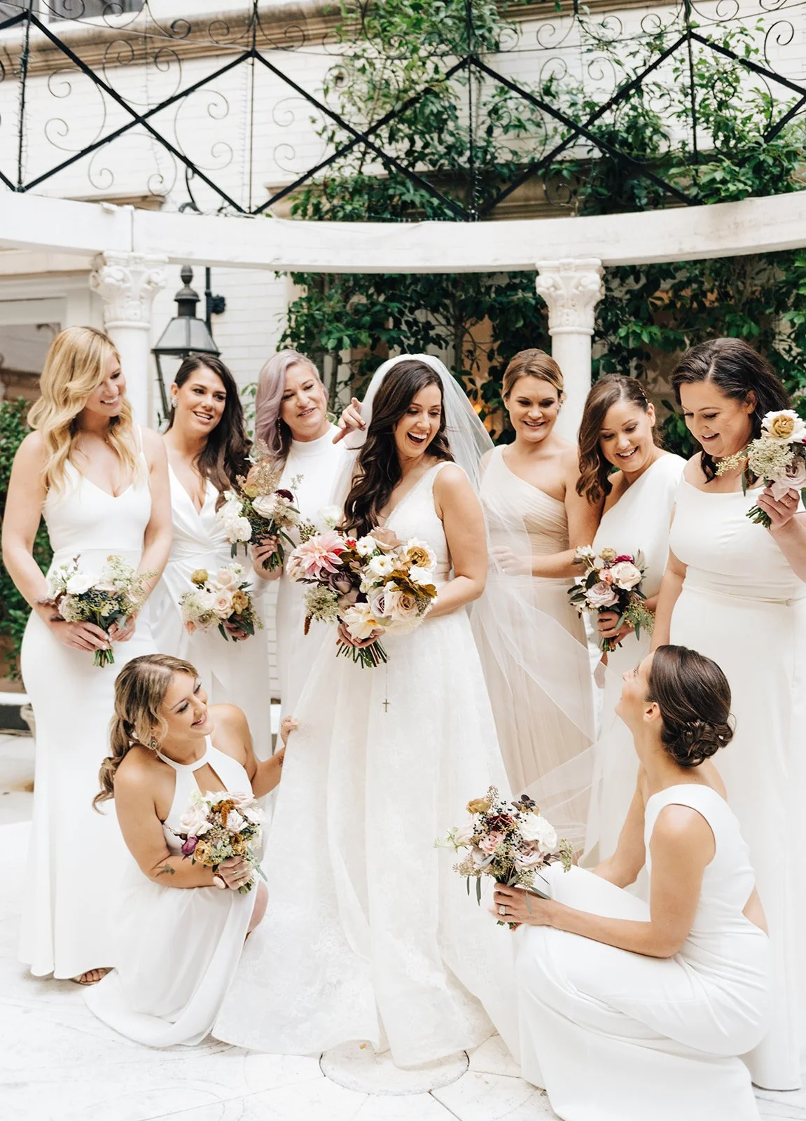 A bride and her bridesmaids, all in white dresses, gathered outdoors on a patio with greenery and ornate columns. The bride is laughing, holding a bouquet, surrounded by her joyful bridesmaids holding flowers.
