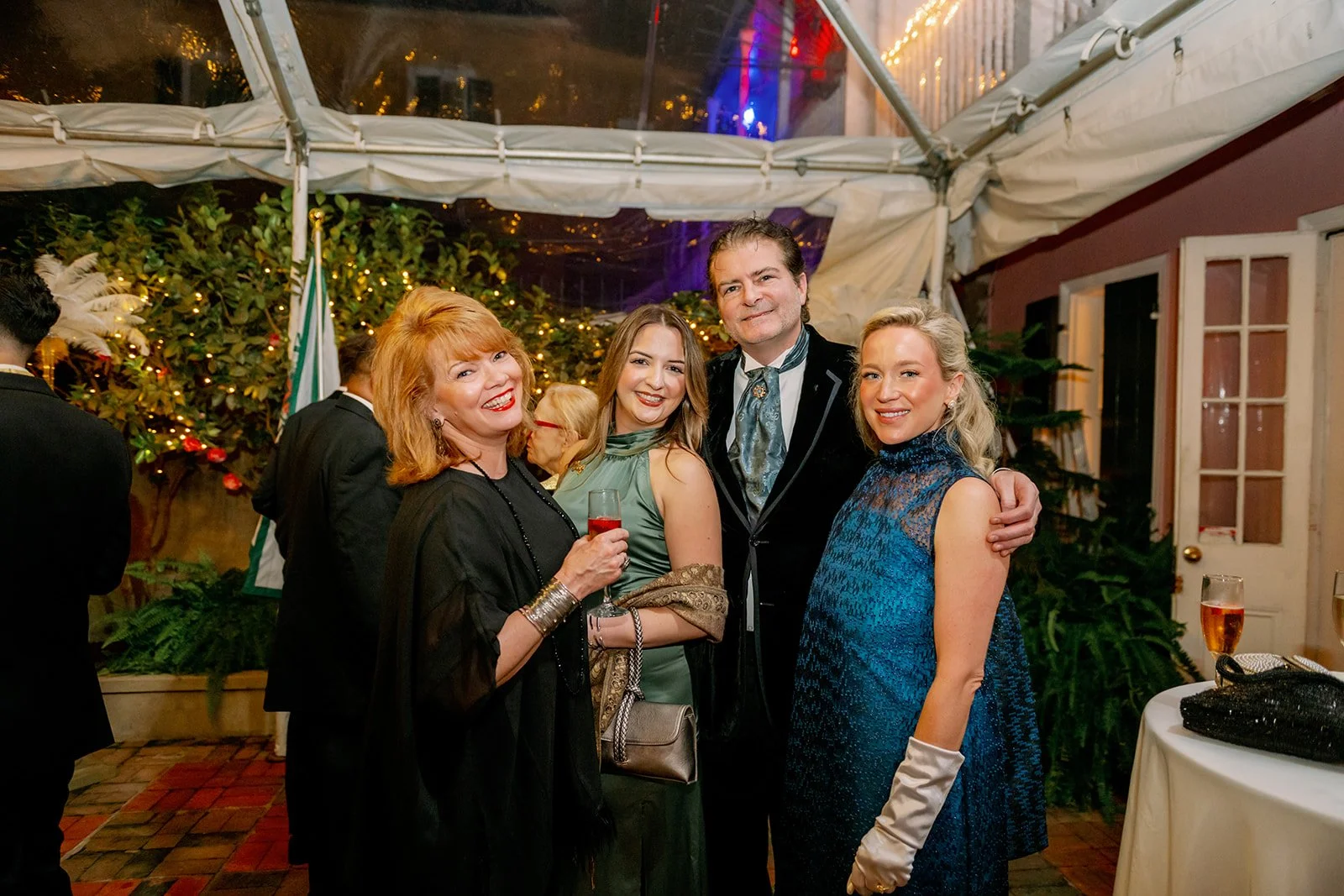 Group of four adults smiling and posing for a photo at a festive gathering, with a decorated outdoor tent, Christmas lights, and greenery in the background, holding drinks.