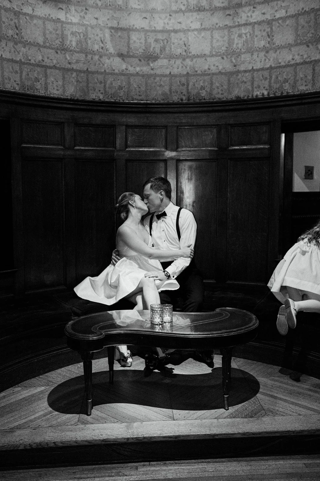 A black-and-white photo of a couple dressed in wedding attire, sharing a kiss while sitting on a bench in a dimly lit room with dark wood paneling.