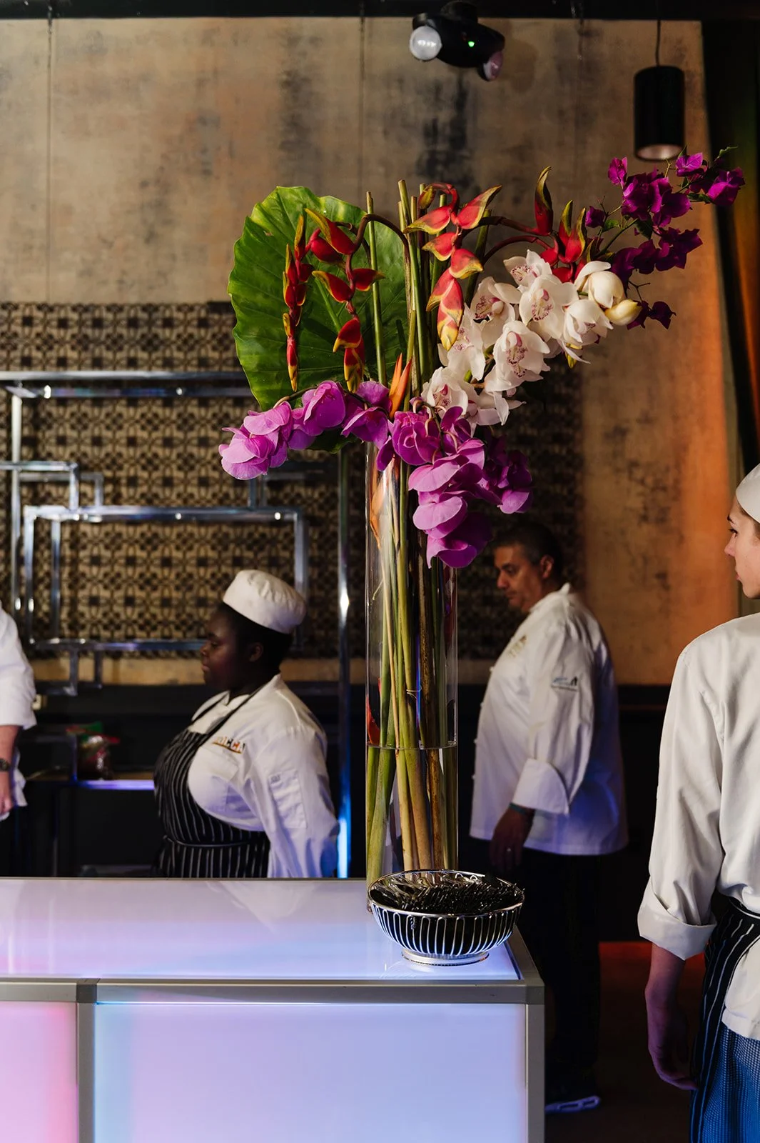 A tall glass vase with colorful orchids and large green leaves on a white countertop in a restaurant or kitchen setting, with chefs in white uniforms and hats in the background.