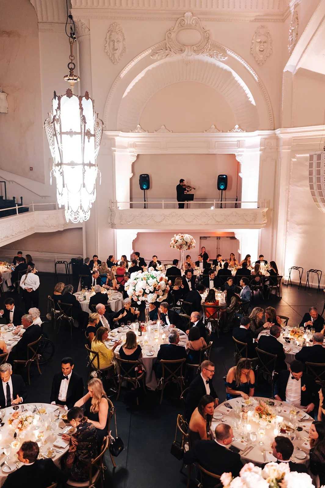 Guests dining at round tables with floral centerpieces in a decorated banquet hall with a violinist on stage