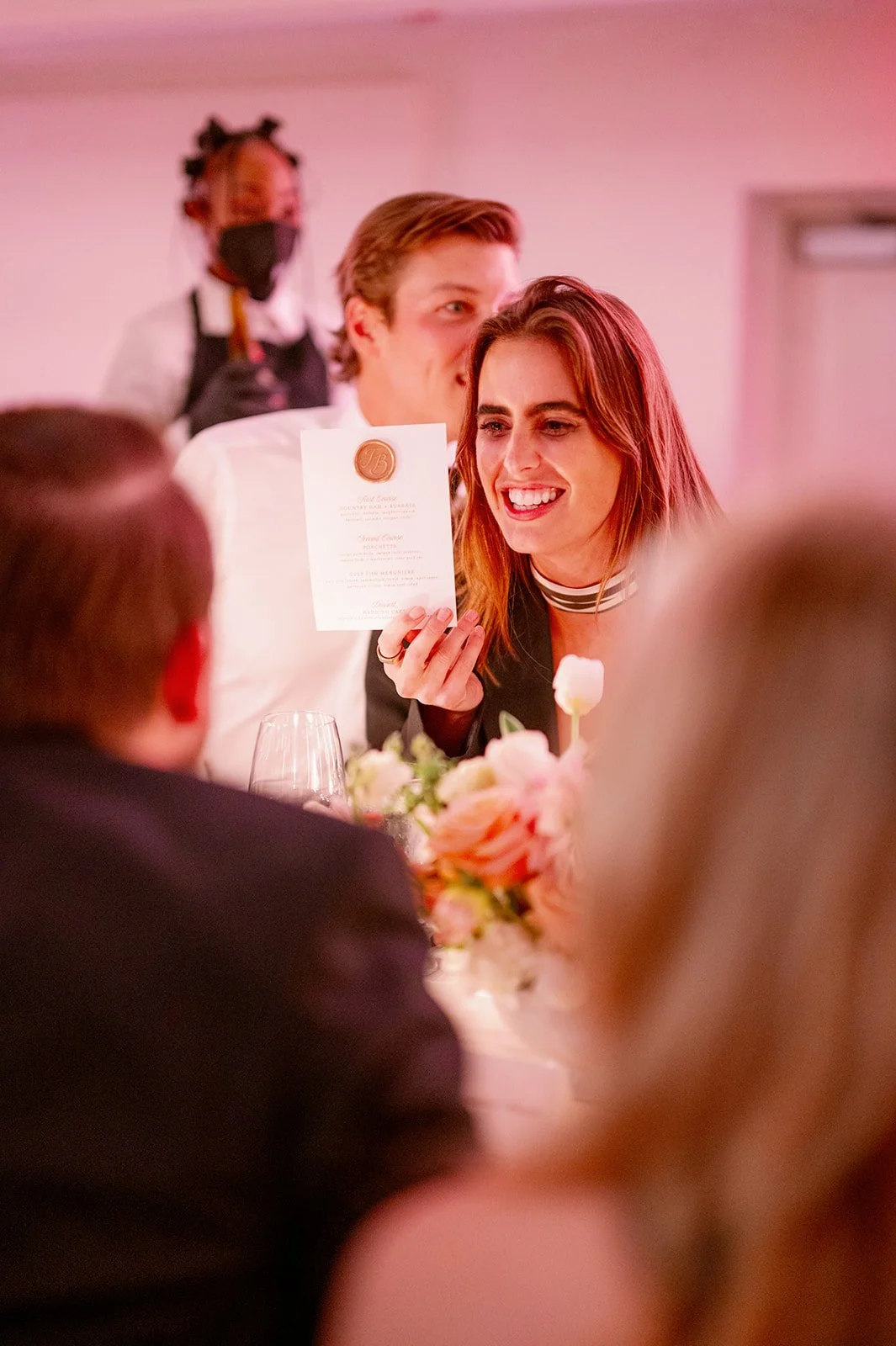 A woman holding a wedding certificate at a wedding reception, smiling and sitting at a table with flowers.