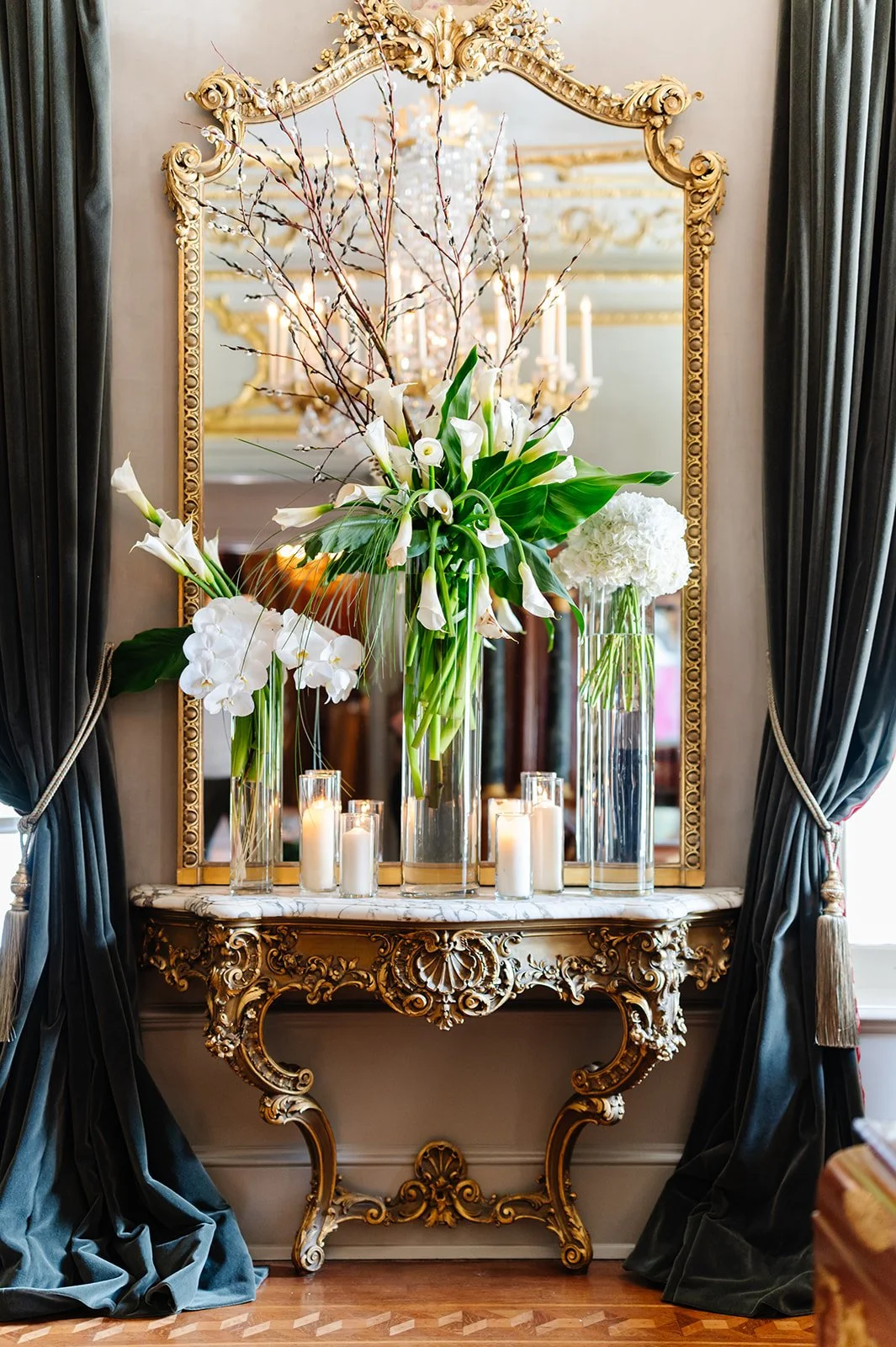 A decorative gold ornate mirror reflecting a chandelier, with a marble-topped gold console table below. The table holds tall glass vases filled with white calla lilies, orchids, and hydrangeas, with candles in glass holders in front.